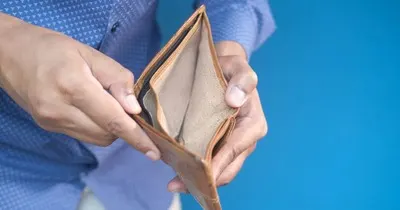 A man in a blue shirt holding an open, empty leather wallet against a blue background, symbolising financial difficulty and over-indebtedness